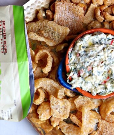 Creamy Kale Artichoke Dip in an orange and blue serving dish, on top of a bed of pork grinds.