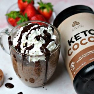 A Keto Chocolate Mug Cake in a glass mug sitting next to a tub of Ancient Nutrition Keto Cocoa