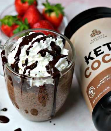A Keto Chocolate Mug Cake in a glass mug sitting next to a tub of Ancient Nutrition Keto Cocoa