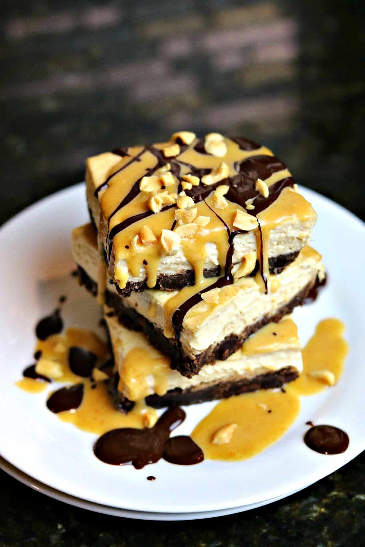 A stack of three Keto Snickers Cheesecake bars, topped with salted caramel drizzle, on a white plate