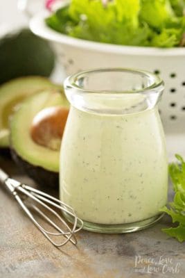 A jar of Creamy Avocado Ranch Dressing with whisk, salad, and avocado