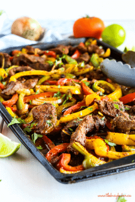 A close-up of steak fajitas on a black sheet pan with sliced peppers, lime wedges and chopped cilantro