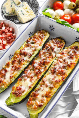 Keto Bacon Zucchini Boats in a white and blue casserole pan with dishes of cheese and vegetables