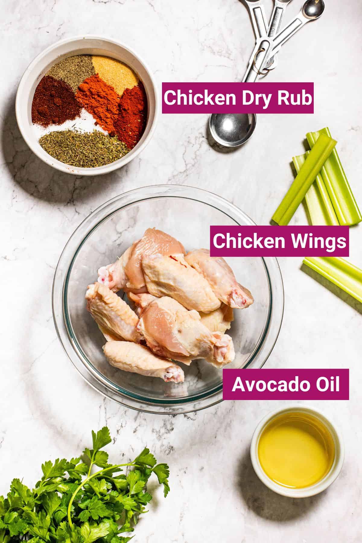 Overhead view of the labeled ingredients needed for spicy dry rub chicken wings: a bowl of raw chicken wings, a bowl of avocado oil, and a bowl of chicken dry rub, next to celery sticks, cilantro, and measuring spoons