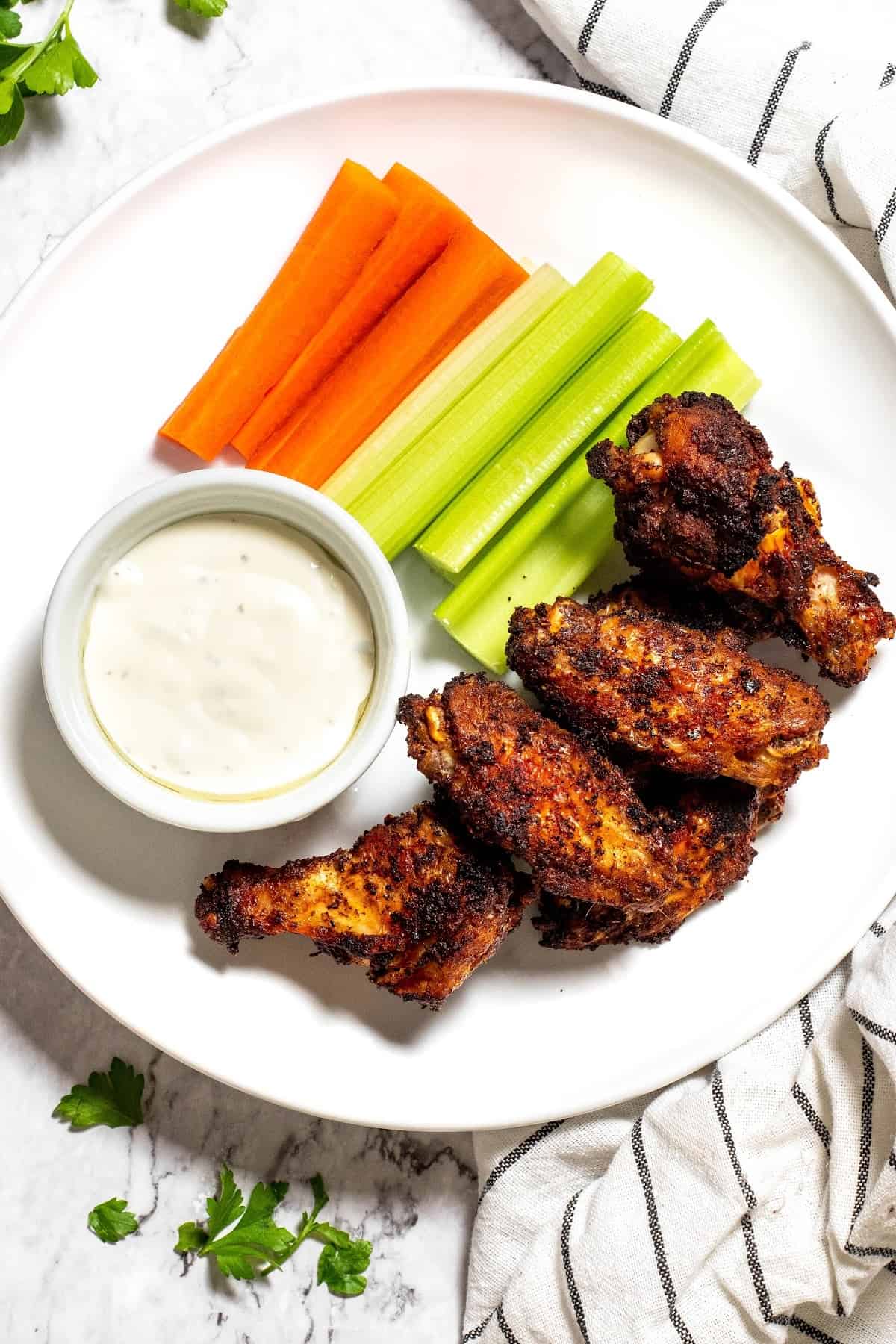 Overhead view of a plate with 4 wings, some celery sticks, some carrot sticks, and a bowl of ranch dressing, next to some parsley and a kitchen towel