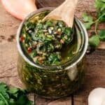chimichurri in a glass jar with a wooden spoon surrounded by parsley