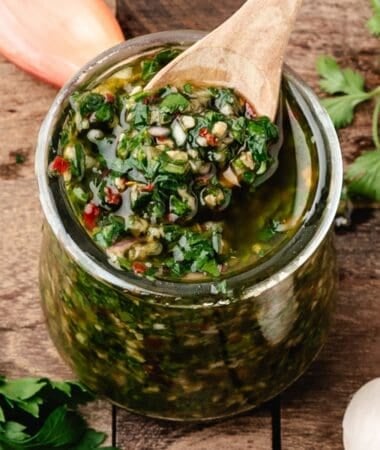 chimichurri in a glass jar with a wooden spoon surrounded by parsley