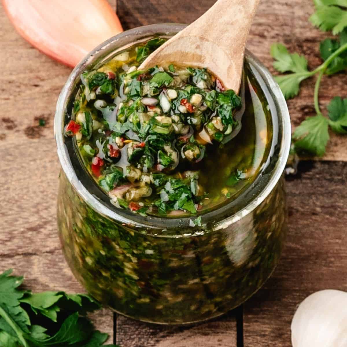 chimichurri in a glass jar with a wooden spoon surrounded by parsley
