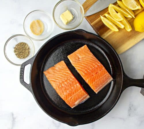 Two Salmon Filets in a pan next to a chopping board with slices of lemon
