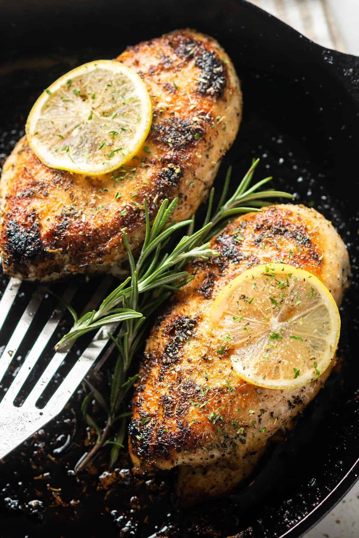 two cooked chicken breasts in a cast iron pan with lemon and rosemary