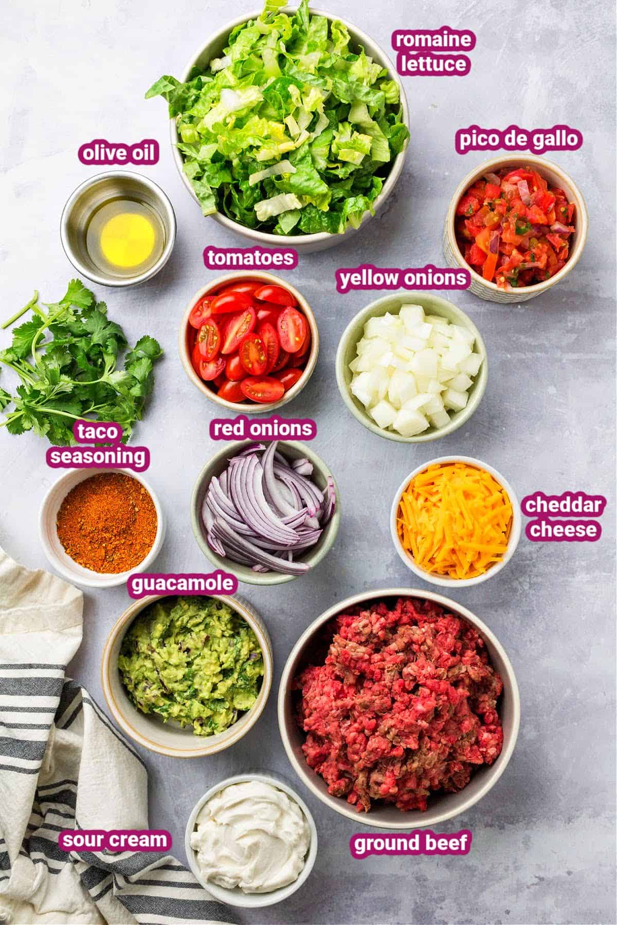 keto taco salad bowl ingredients like lettuce, salsa, taco seasoning, ground beef, red onions, tomato, cheddar cheese, yellow onions, guacamole, olive oil, and sour cream