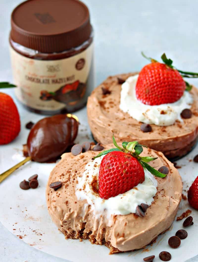 two keto no bake chocolate cheesecakes on a parchment paper with chocolate hazelnut spread on a spoon