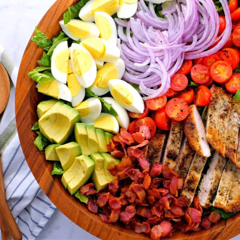a close-up of a Chicken Cobb Salad with eggs, avocado, bacon, red onions, and cast iron skillet chicken breast in a brown bowl