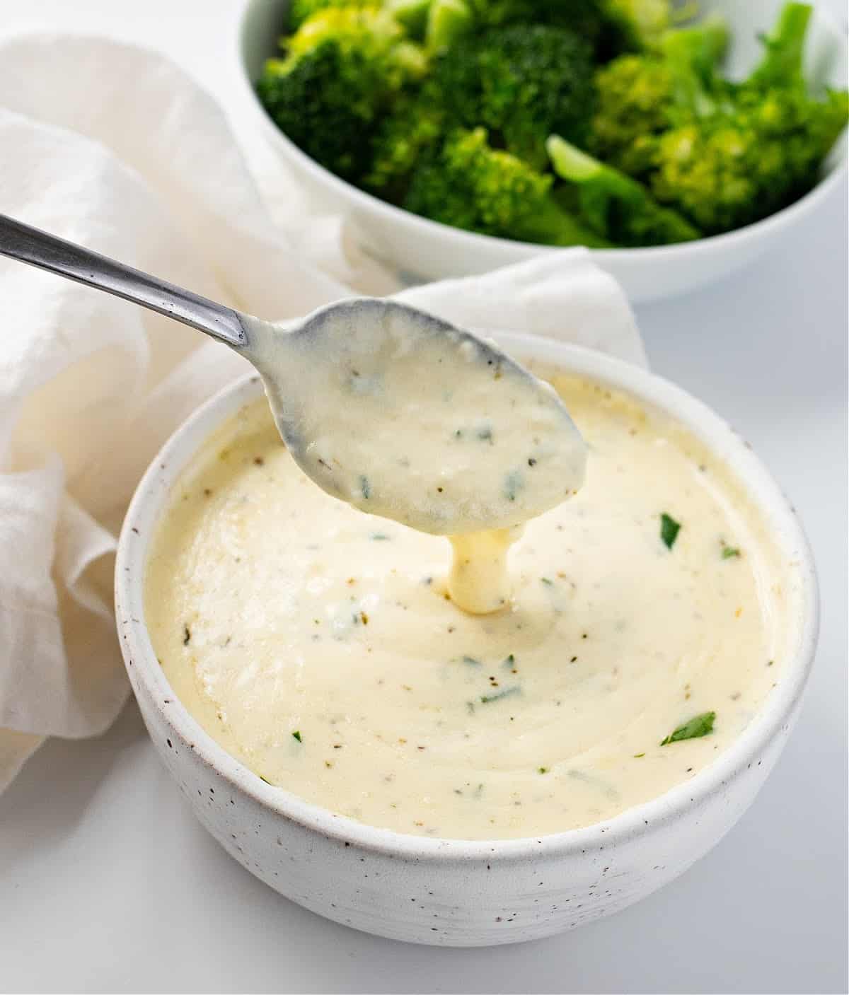 Keto Alfredo sauce in a bowl with a spoon and broccoli in the back