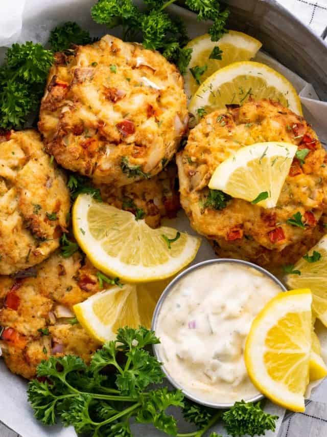 a close-up of six air fryer crab cakes in a metal bowl with lemon, dill, parsley, and homemade tartar sauce