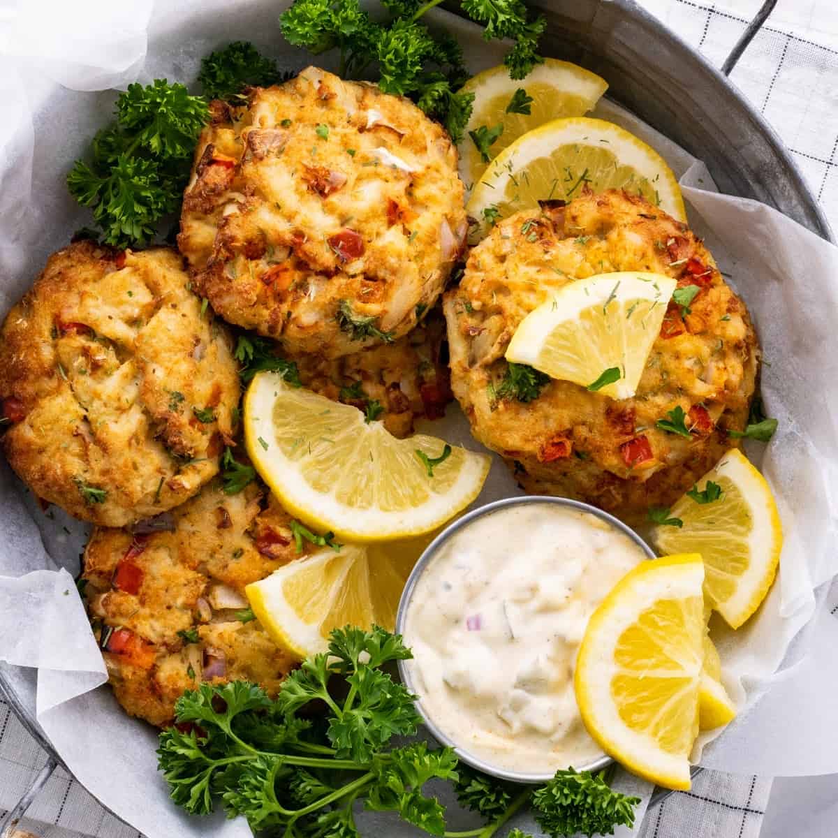 a close-up of six air fryer crab cakes in a metal bowl with lemon, dill, parsley, and homemade tartar sauce