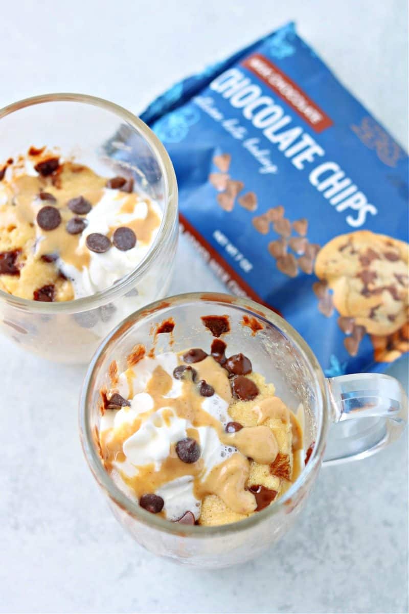 two keto peanut butter mug cakes in glass mugs topped with whipped cream, keto milk chocolate, and peanut butter