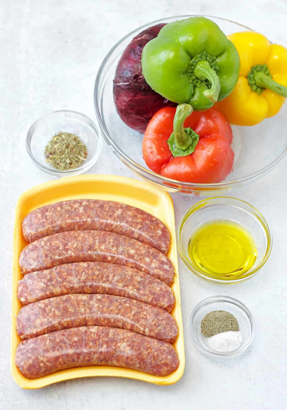 ingredients needed to make air fryer Italian sausages like raw pork sausage, Italian seasoning herb blend, olive oil, salt, black pepper, red onion, and bell peppers in separate bowls.