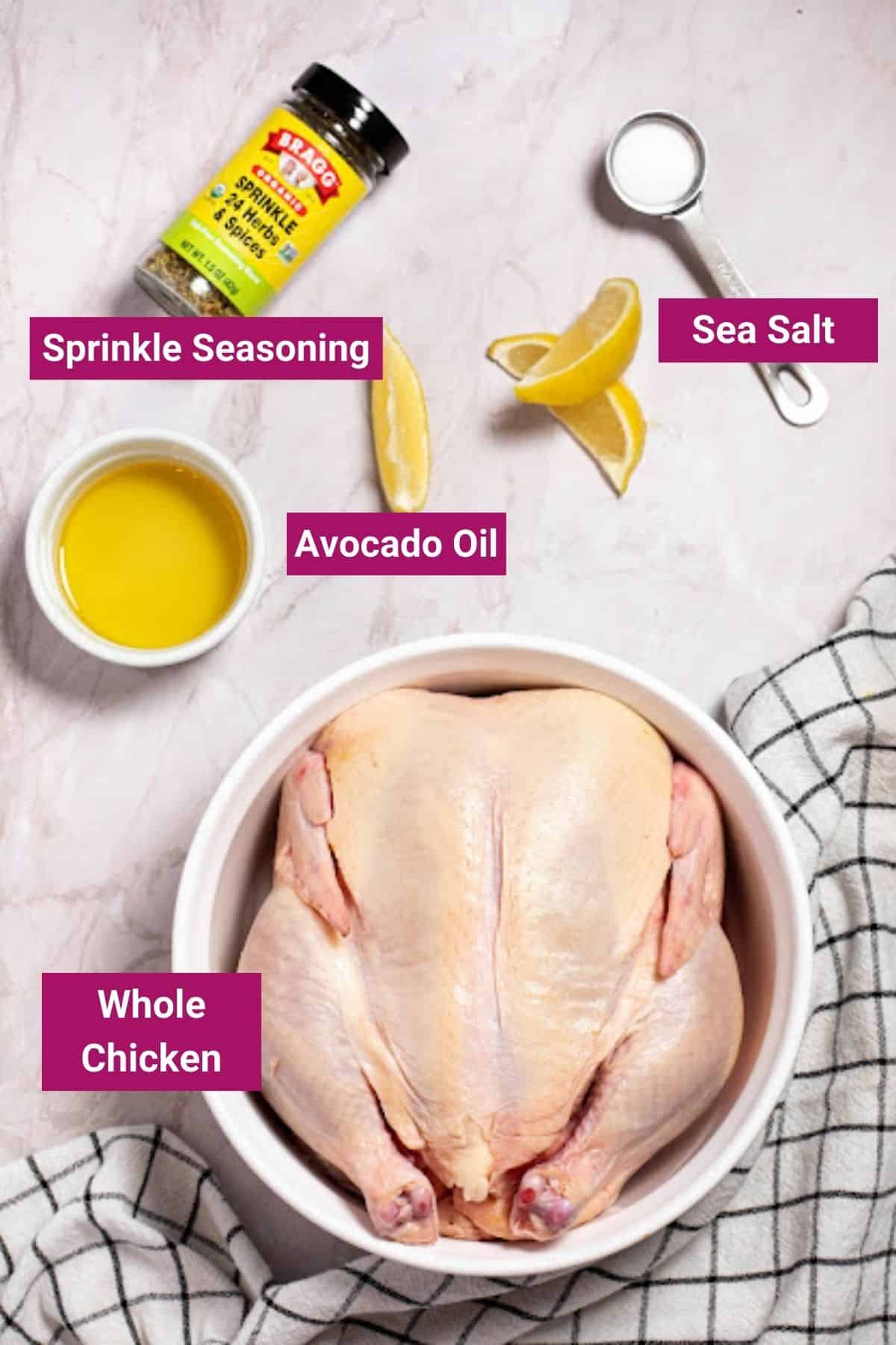 Overhead view of the labeled ingredients for air fryer roasted chicken: a bowl with a whole, raw chicken, a bowl of avocado oil, a jar of sprinkle seasoning, and a measuring spoon of sea salt, next to some lemon slices