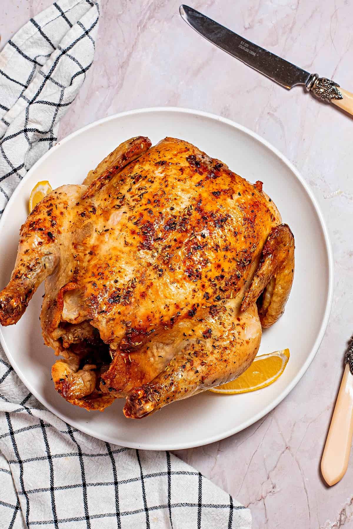 Overhead view of a roast chicken on a plate with a lemon slice, next to a kitchen towel and a knife