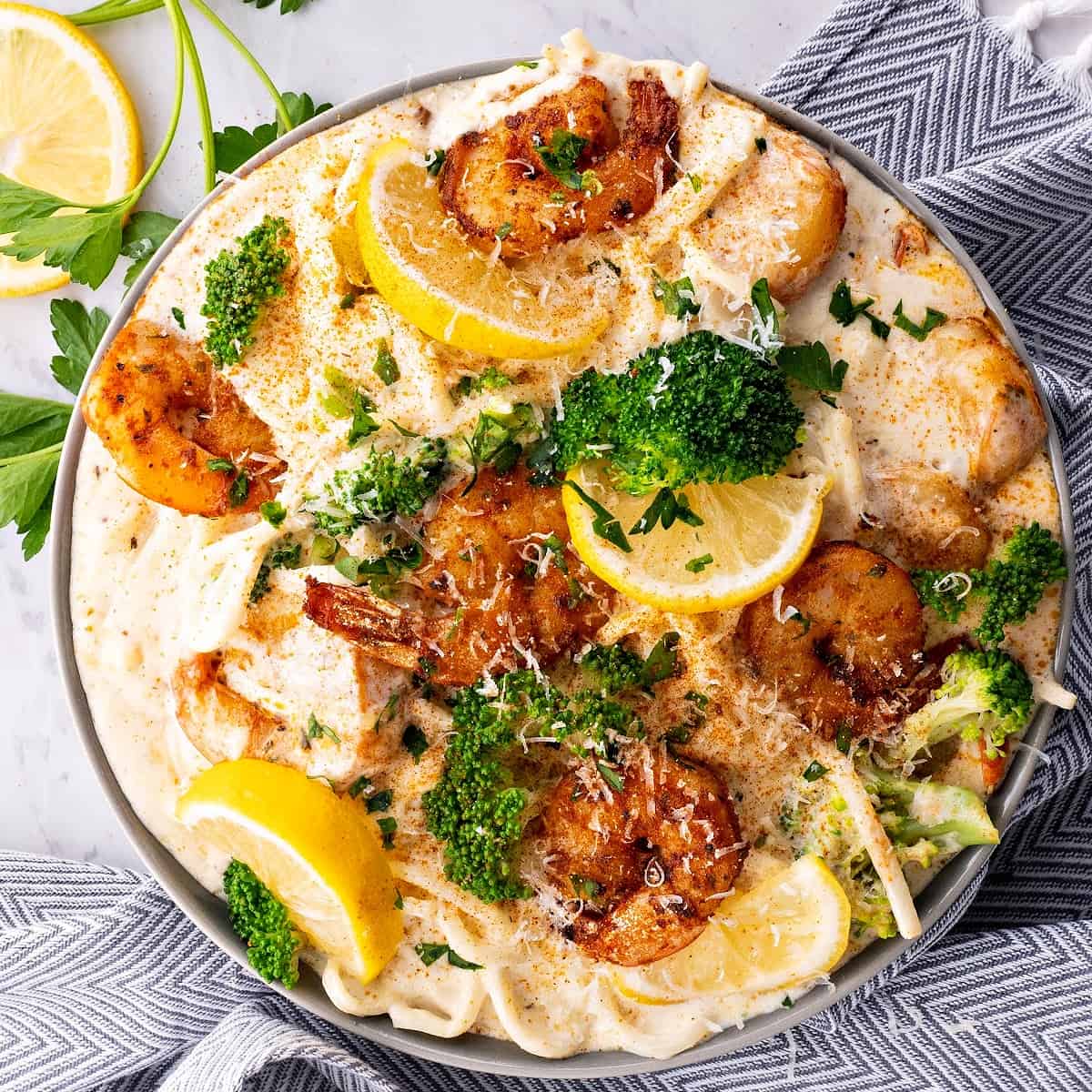 close up of cajun shrimp alfredo pasta in a bowl with broccoli, parsley and lemon wedges