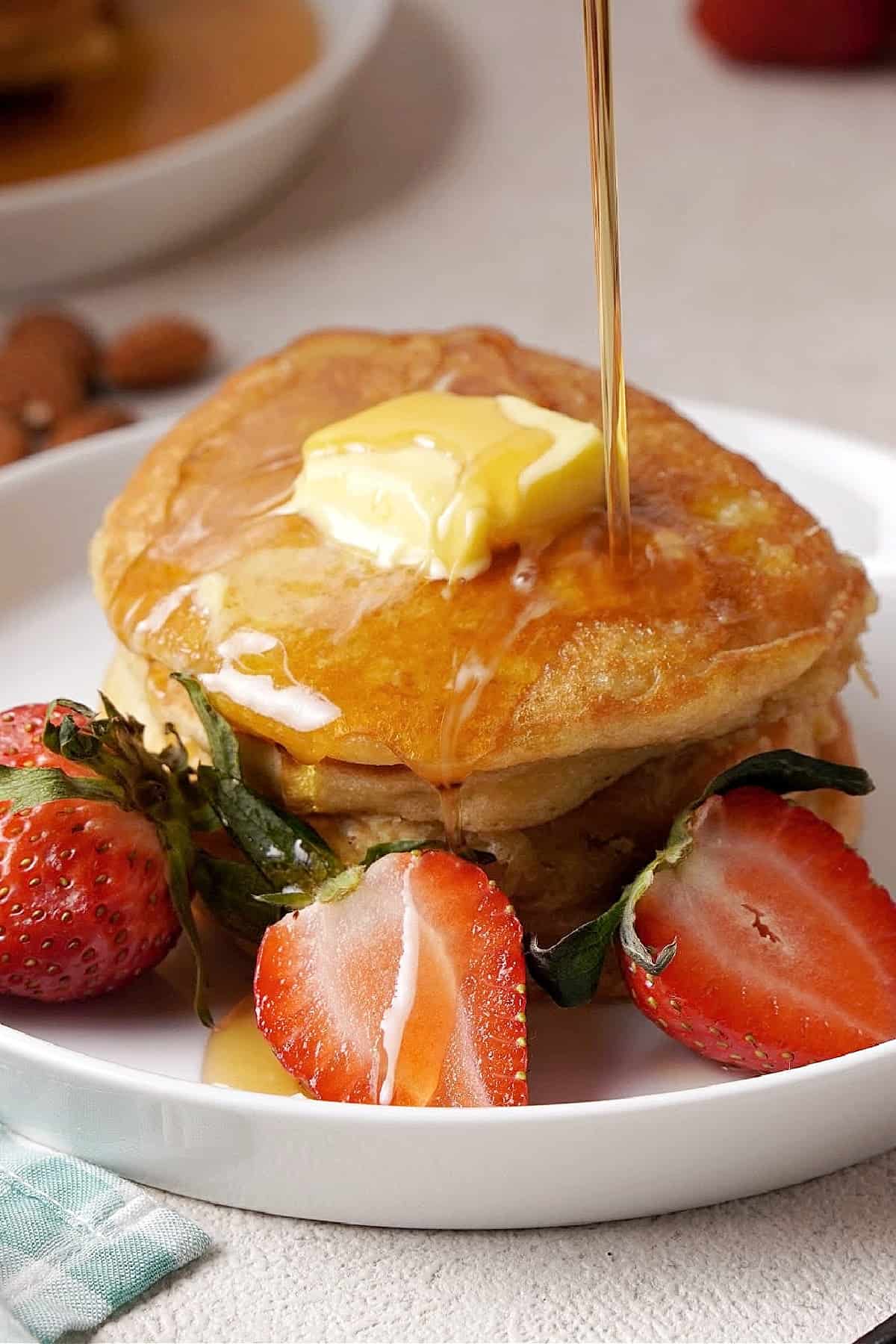 Maple syrup being poured over a stack of pancakes with butter on top, with three strawberries on the plate 