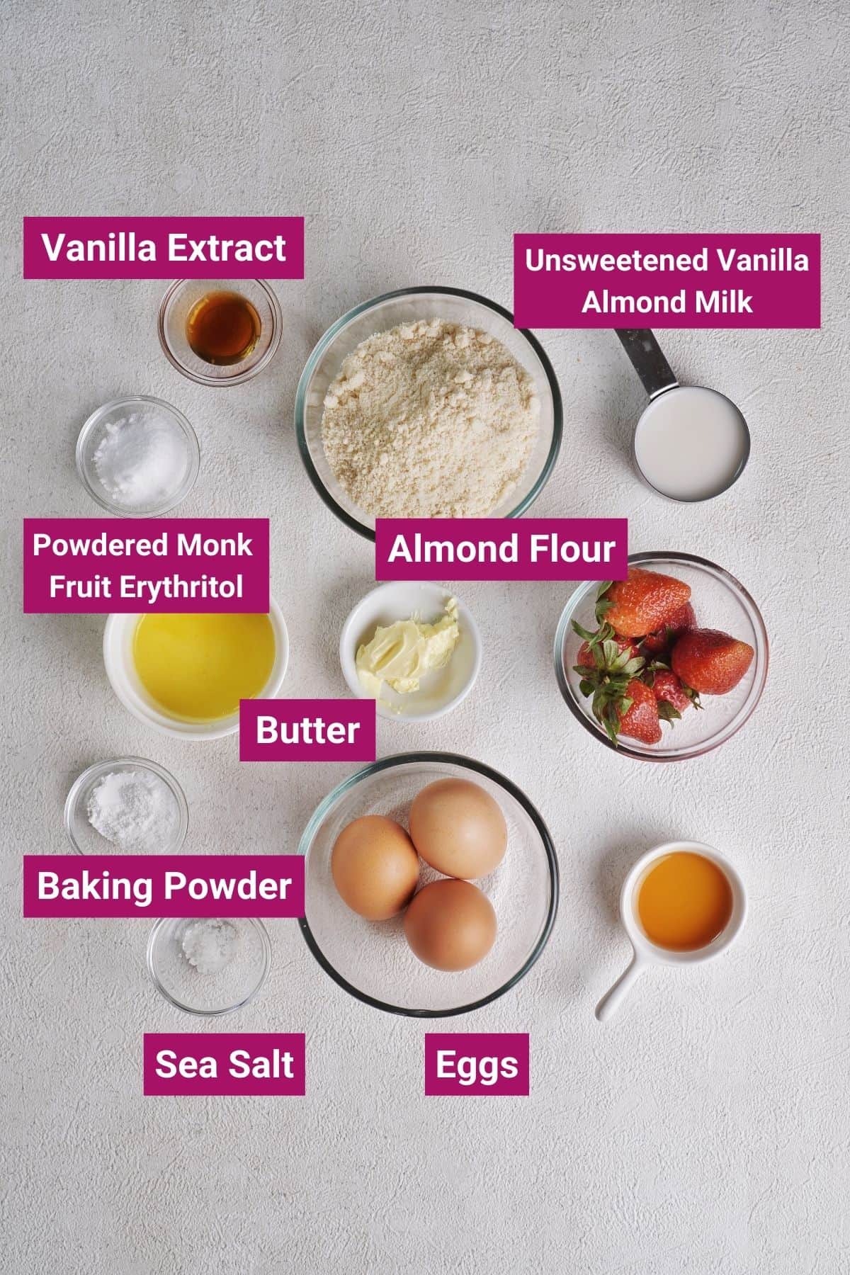 Overhead view of the labeled ingredients needed for keto pancakes: a measuring cup of unsweetened vanilla almond milk, a bowl of almond flour, a bowl of vanilla extract, a bowl of powdered monk fruit erythritol, a bowl of butter, a bowl of baking powder, a bowl of sea salt, and a bowl of eggs, next to a bowl of strawberries and some maple syrup. 