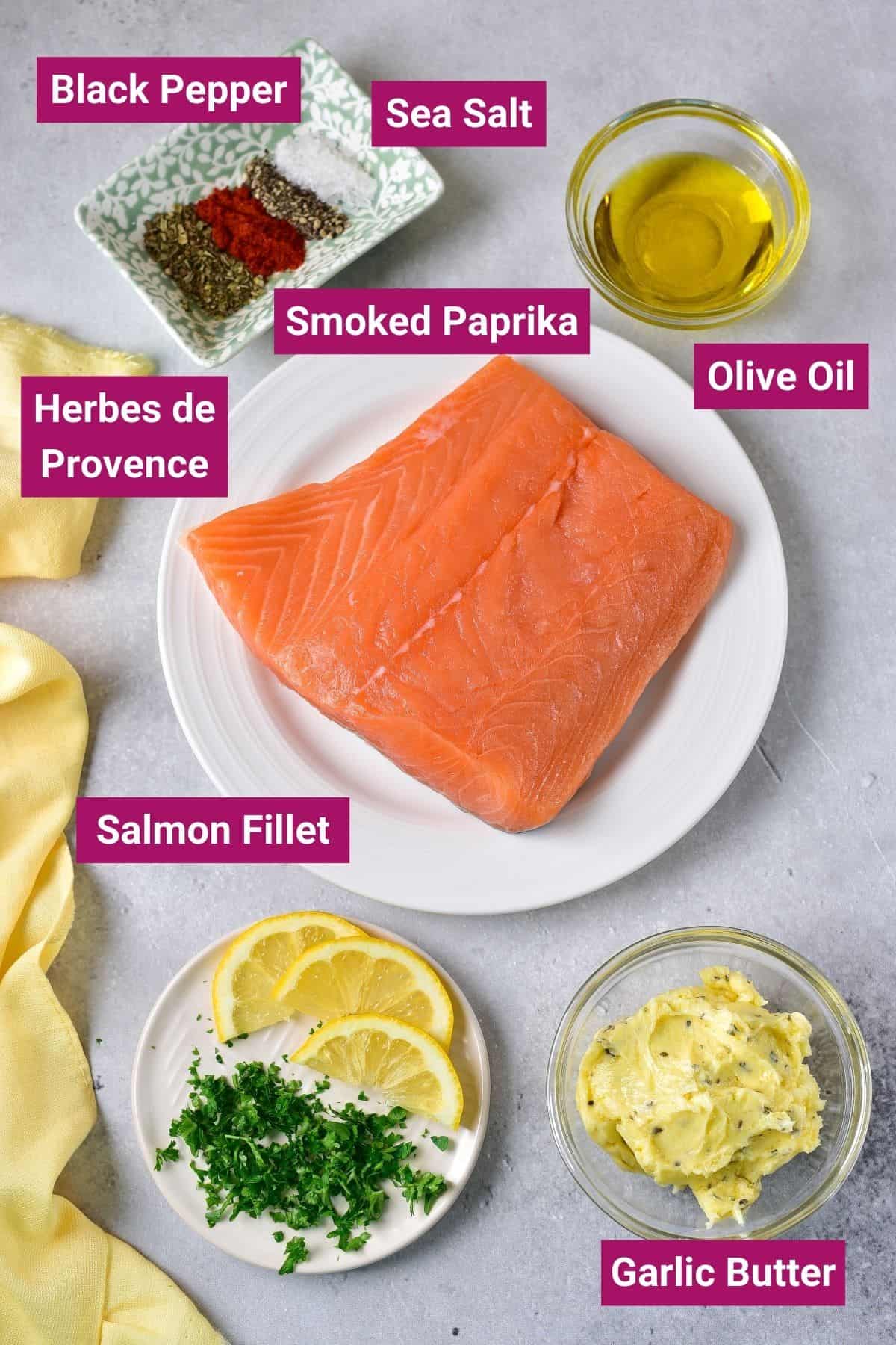 Overhead view of the labeled ingredients needed for air fryer salmon fillets: A plate with a salmon filed, a ramekin of olive oil, a dish of sea salt, black pepper, smoked paprika, and Herbes de Provence, a ramekin of garlic butter, and a plate of lemon slices and chopped parsley