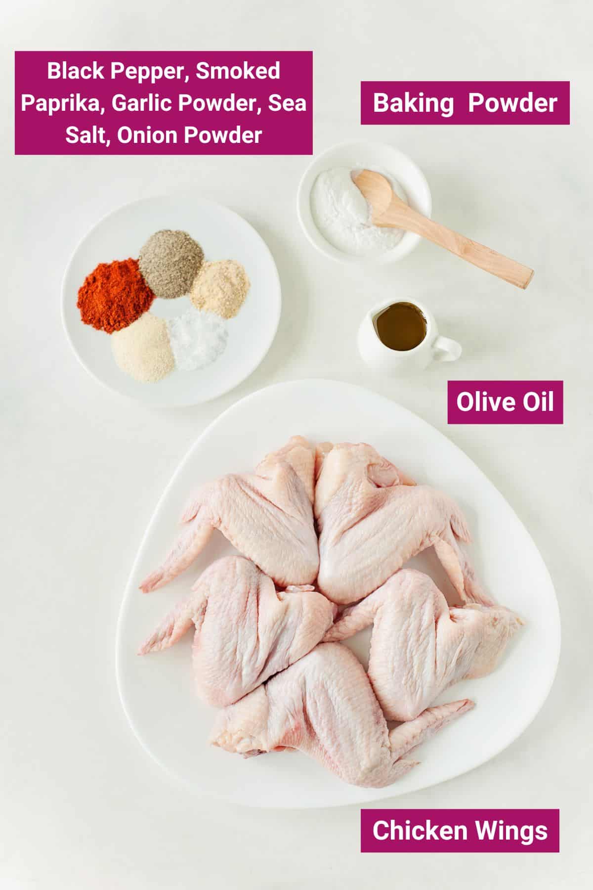 Overhead view of the labeled ingredients needed for air fried wings:  a plate of raw chicken wings, a jug of olive oil, a bowl of baking powder with a spoon, and a plate with black pepper, smoked paprika, garlic powder, sea salt, and onion powder