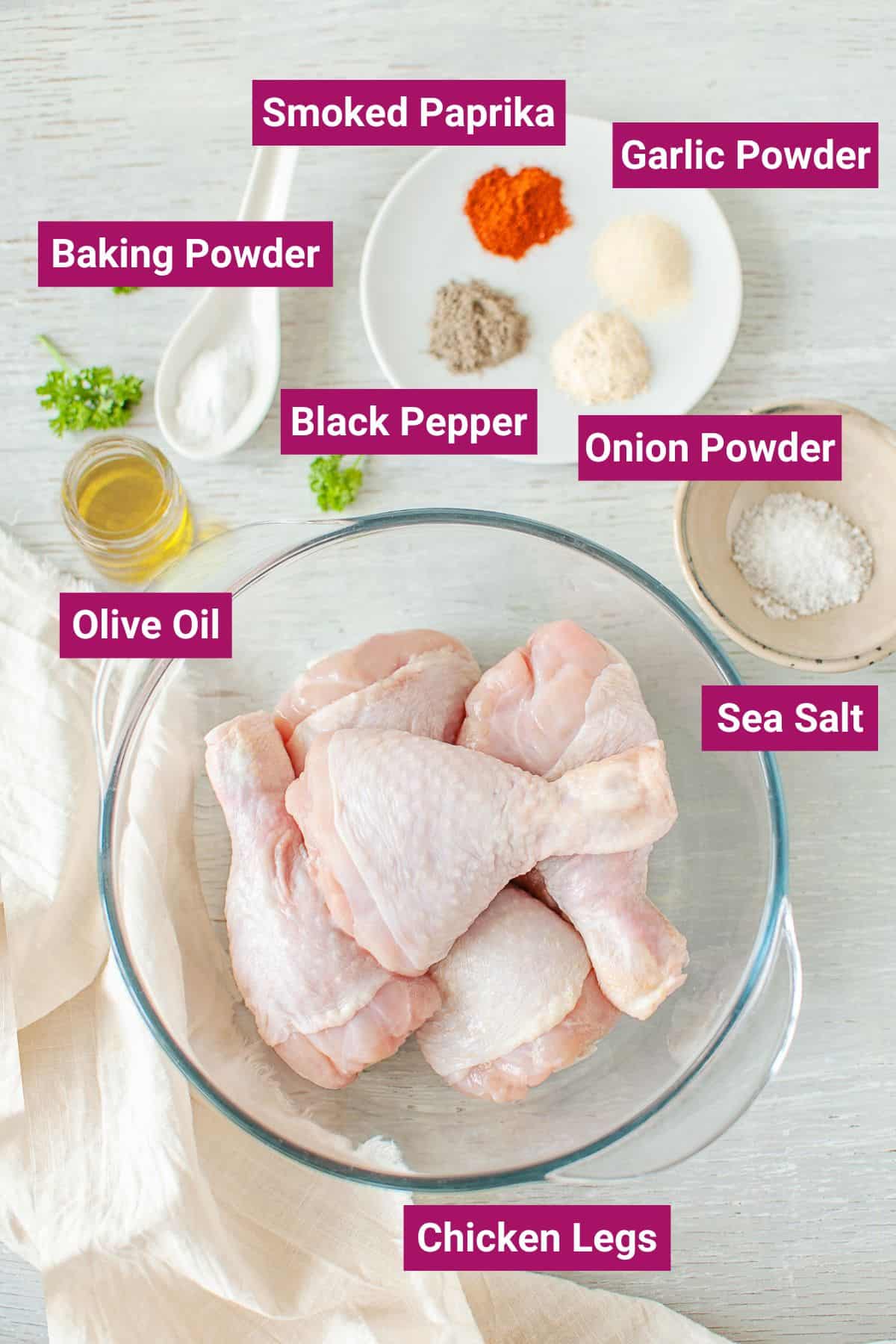 Overhead view of the labeled ingredients for air fryer chicken: a bowl of raw chicken legs, a bowl of sea salt, a plate with onion powder, black pepper, garlic powder, and smoked paprika, a spoon with baking powder, and a jar with olive oil