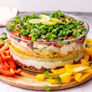 Close up of seven layer dip in a glass serving bowl with lime slices on top, with red bell pepper slices to the left and yellow bell pepper slices to the right