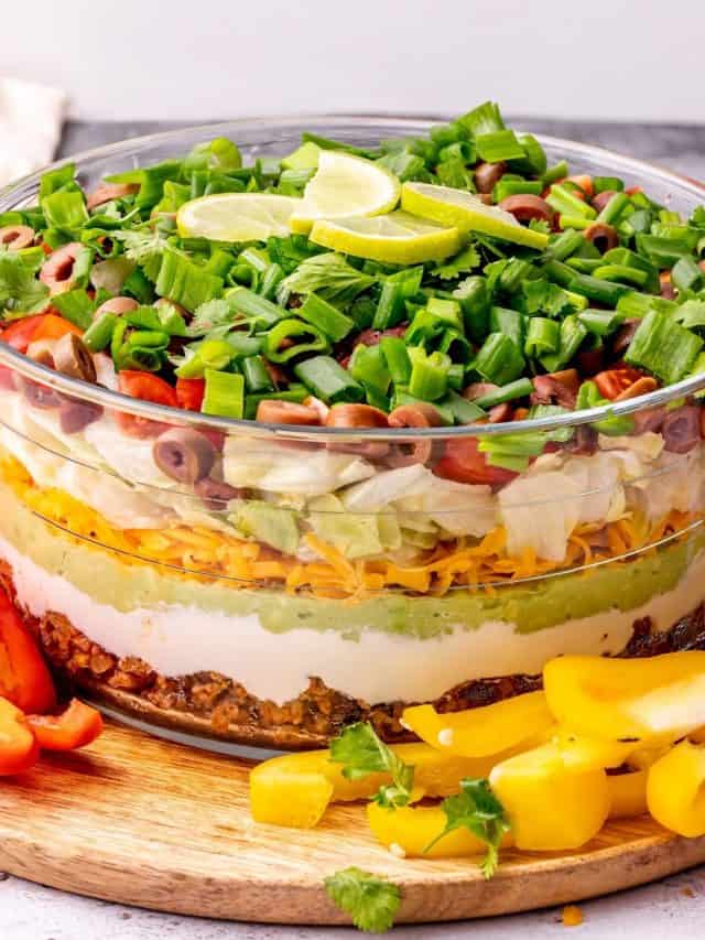 Close up of seven layer dip in a glass serving bowl with lime slices on top, with red bell pepper slices to the left and yellow bell pepper slices to the right