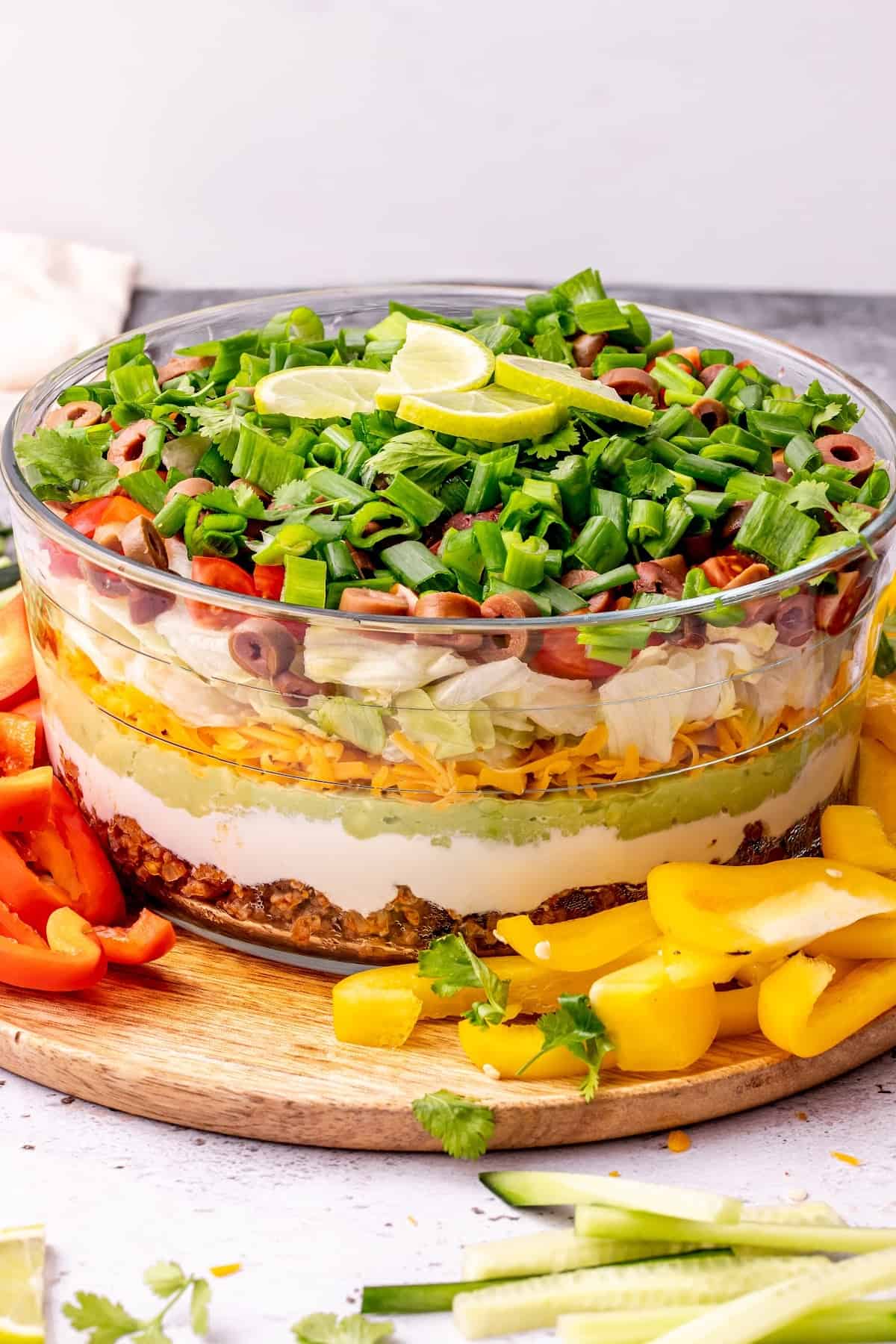 A serving dish of 7 layer dip with lime slices on top, with red bell pepper slices to the left and yellow bell pepper slices to the right