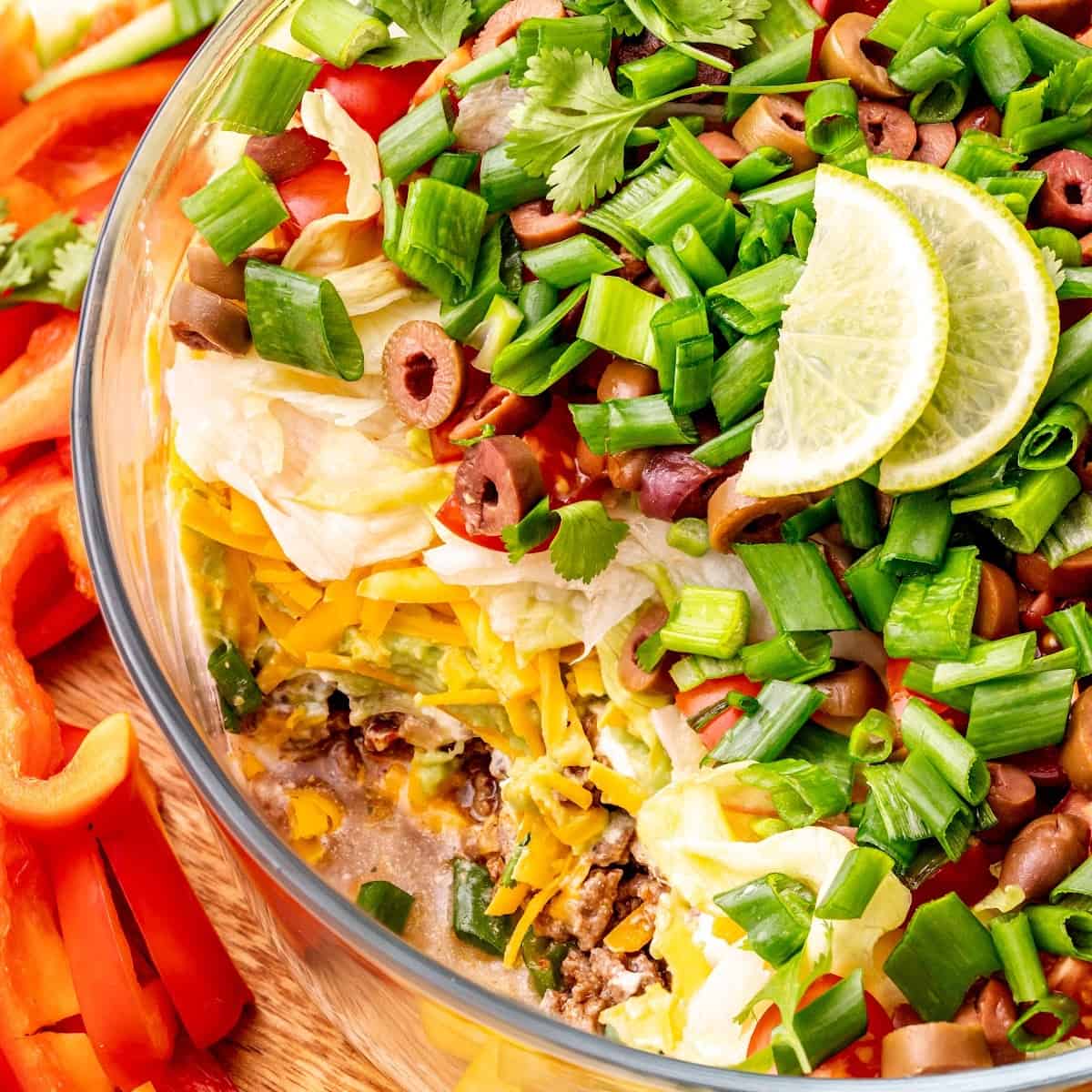 Close up of a serving dish of 7 layer dip topped with lime slices and a few scoops taken out, surrounded by sliced bell peppers