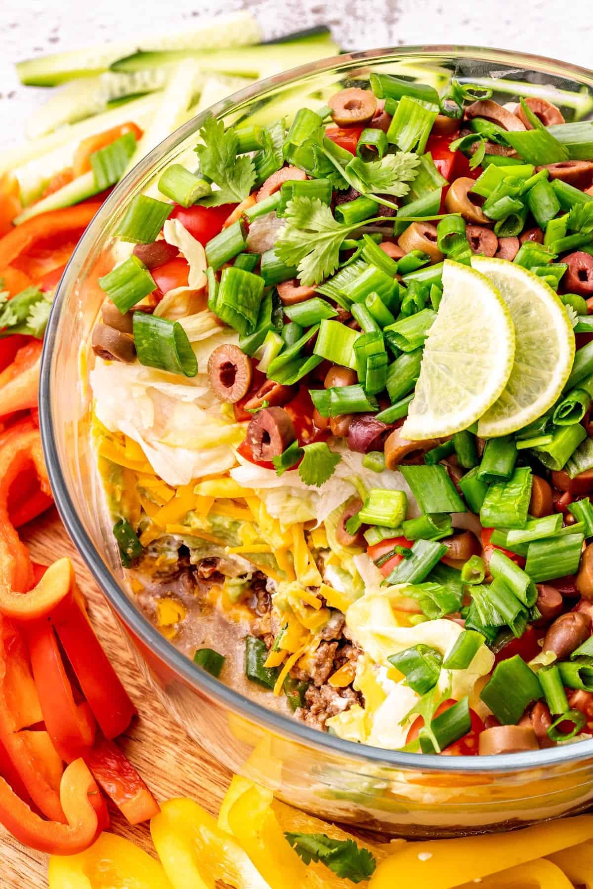 Overhead view of a bowl of 7 layer dip, topped with lime slices, with some scoops taken out, surrounded by bell pepper slices