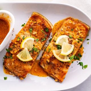 Overhead view of two pieces of salmon on a plate, covered in sauce, parsley, and lemon slices, next to a bowl of sauce