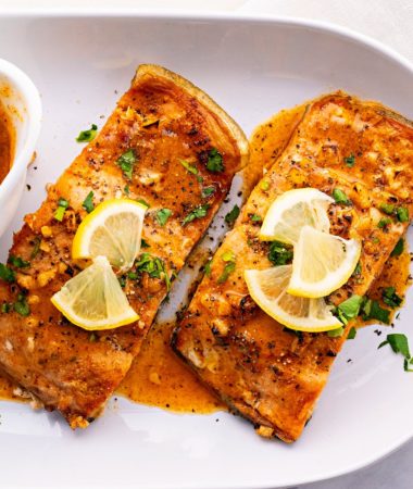 Overhead view of two pieces of salmon on a plate, covered in sauce, parsley, and lemon slices, next to a bowl of sauce