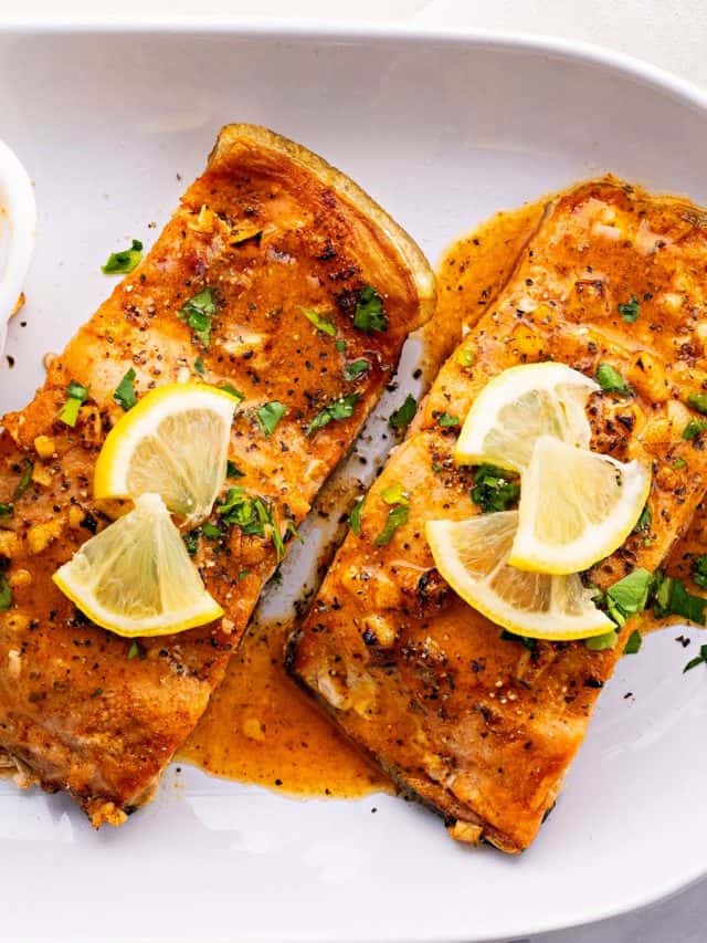 Overhead view of two pieces of salmon on a plate, covered in sauce, parsley, and lemon slices, next to a bowl of sauce