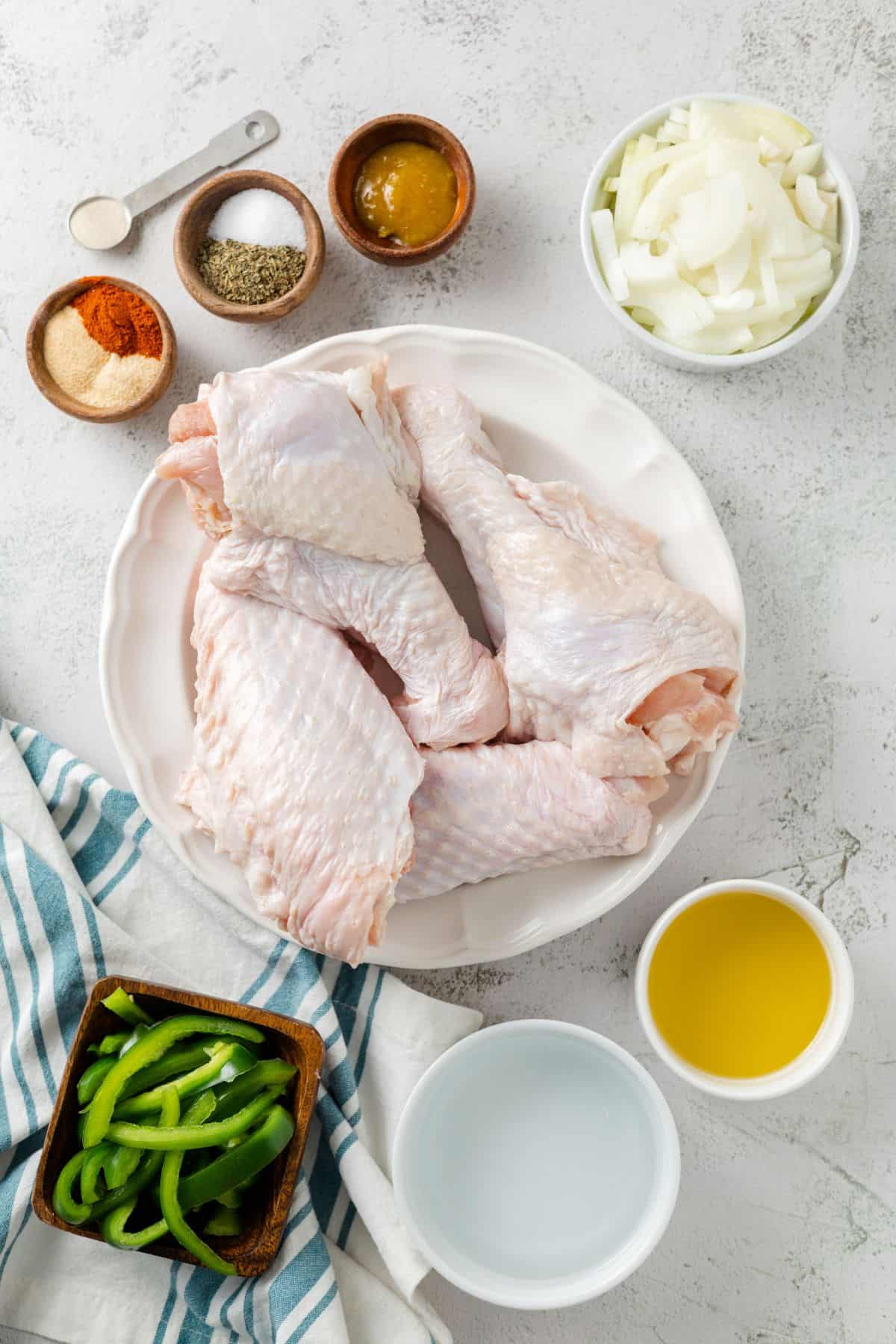 Overhead view of the ingredients needed for crockpot turkey wings: a plate of turkey wings, a bowl of green bell peppers, a bowl of onions, a bowl of oil, a bowl of water, a bowl of broth paste, a bowl of salt and pepper, and a bowl of spices