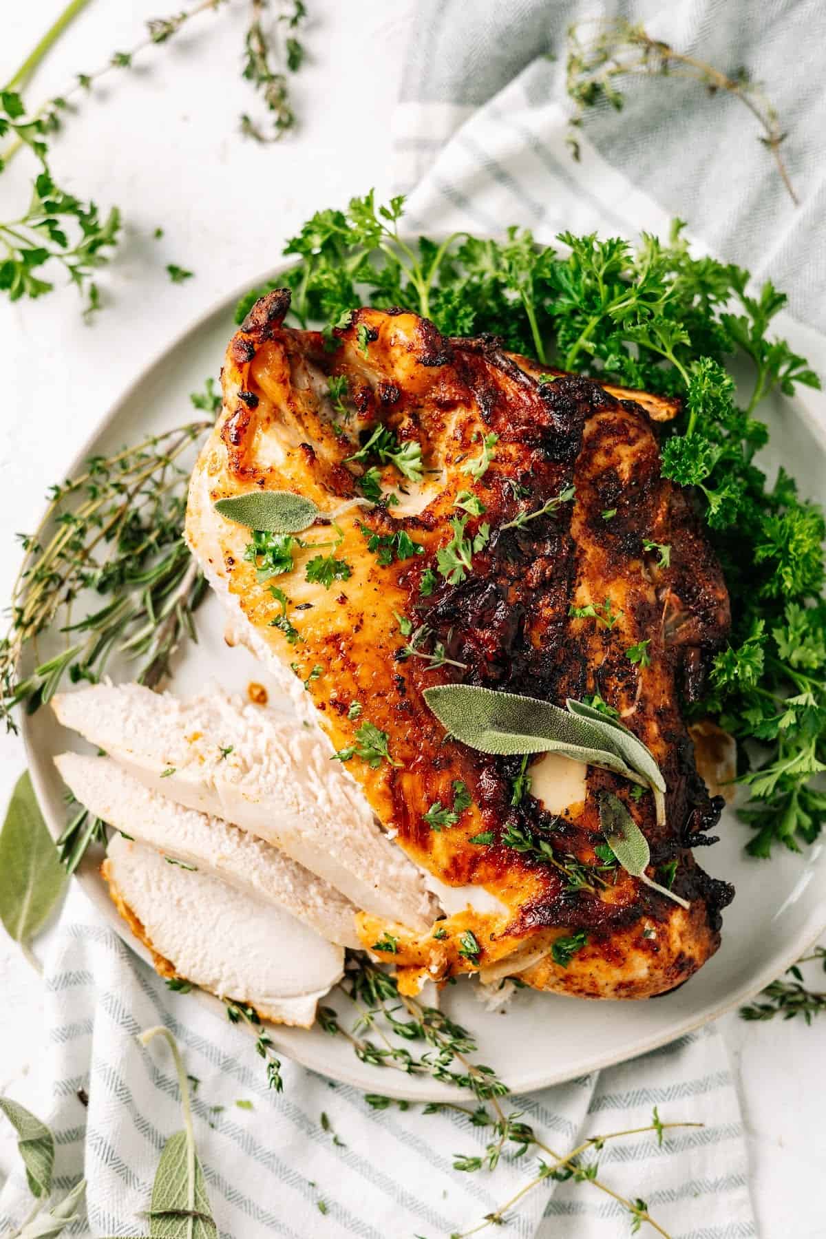 Overhead view of a turkey breast on a plate, with the end cut into slices, covered in herbs
