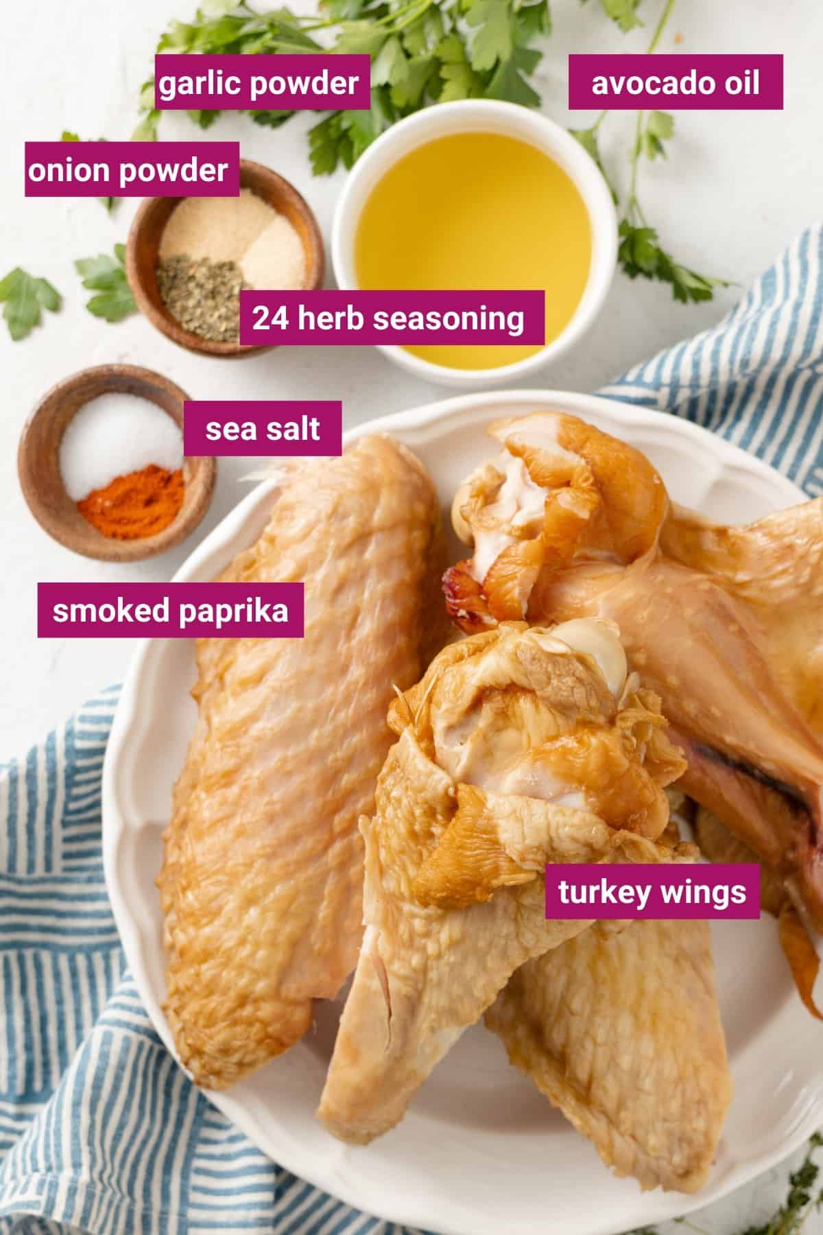 Overhead view of the labeled ingredients for air fryer turkey wings: a plate full of turkey wings, a bowl of sea salt and smoked paprika, a bowl of onion powder, garlic powder, and 24 herb seasoning, and a bowl of avocado oil