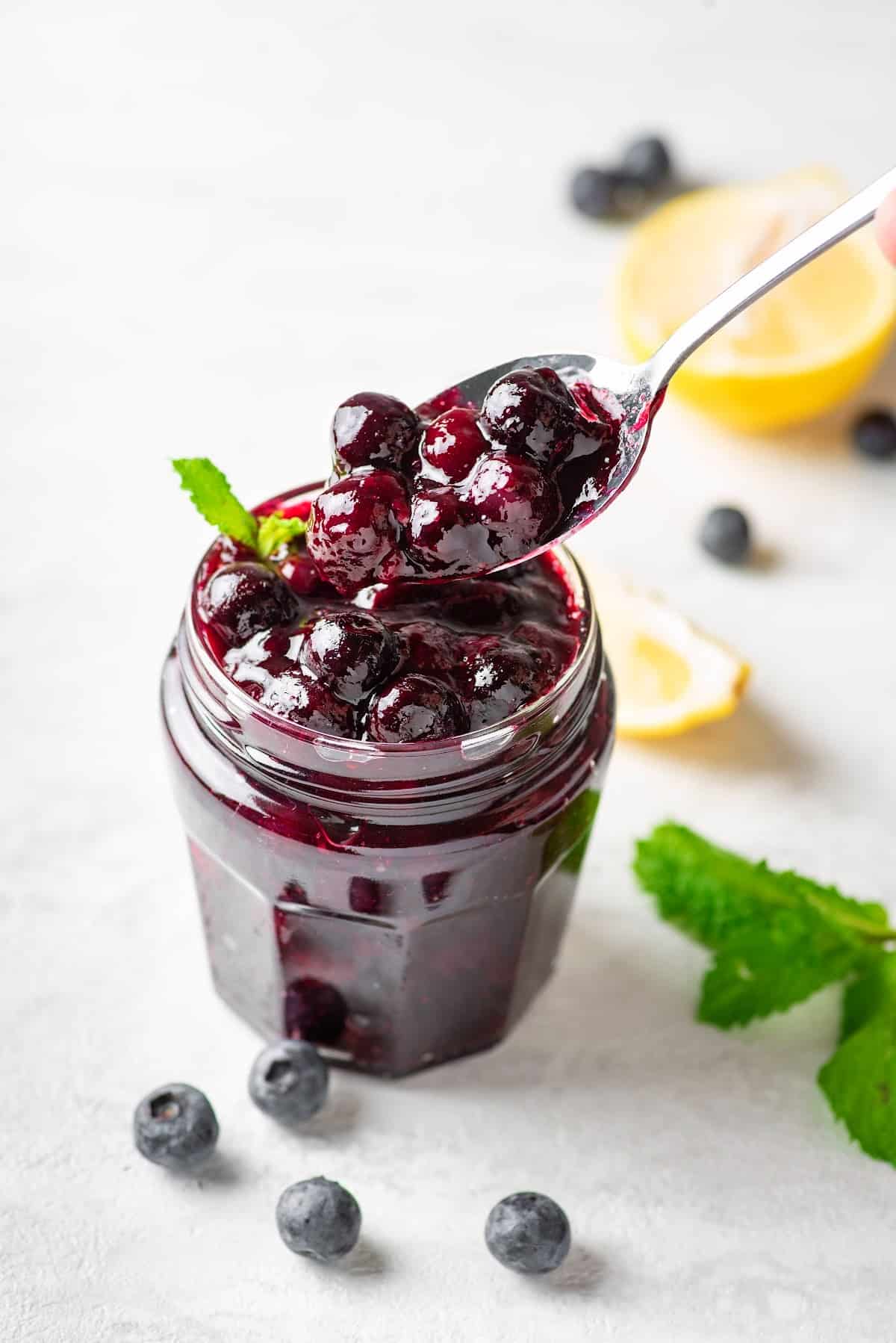 keto blueberry sauce recipe in a glass jar with fresh blueberries, lemon wedges and mint surrounding it