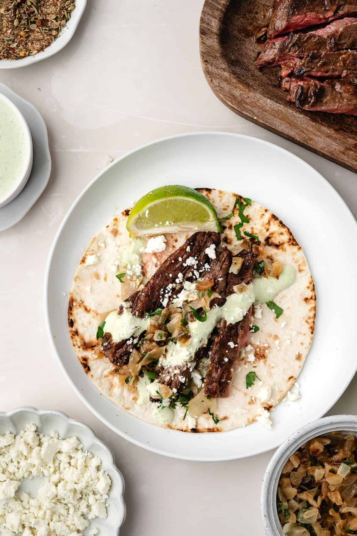 Steak tacos served on a plate with creamy avocado sauce, cotija cheese, and fresh lime slices