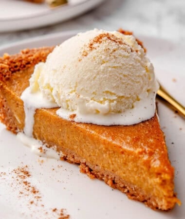 a slice of pumpkin pie with graham cracker crust on a plate topped with a scoop of vanilla ice cream and some pumpkin pie spice sprinkles
