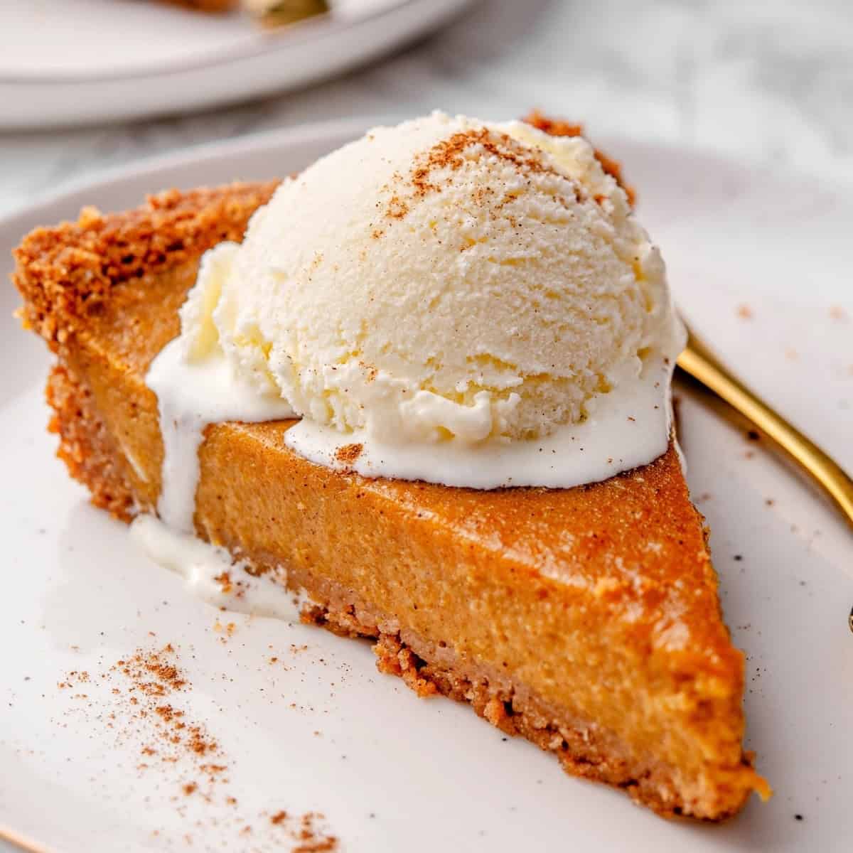a slice of pumpkin pie with graham cracker crust on a plate topped with a scoop of vanilla ice cream and some pumpkin pie spice sprinkles