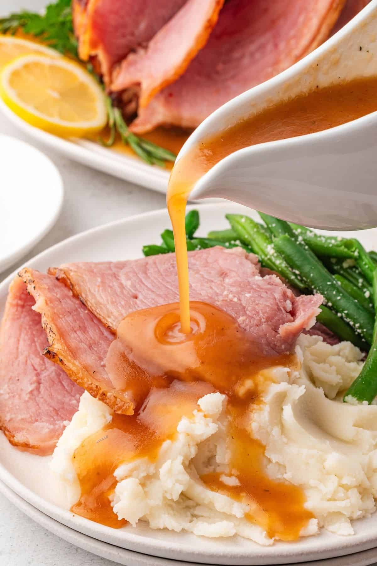 sliced crockpot ham on a plate with green beans and mashed potatoes with ham glaze being poured on top