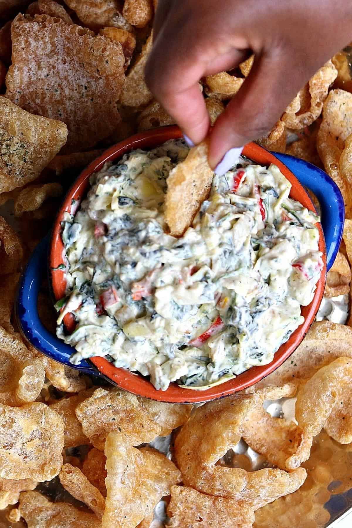 kale artichoke dip in a blue bowl being dipped out by Davinah with a pork rind