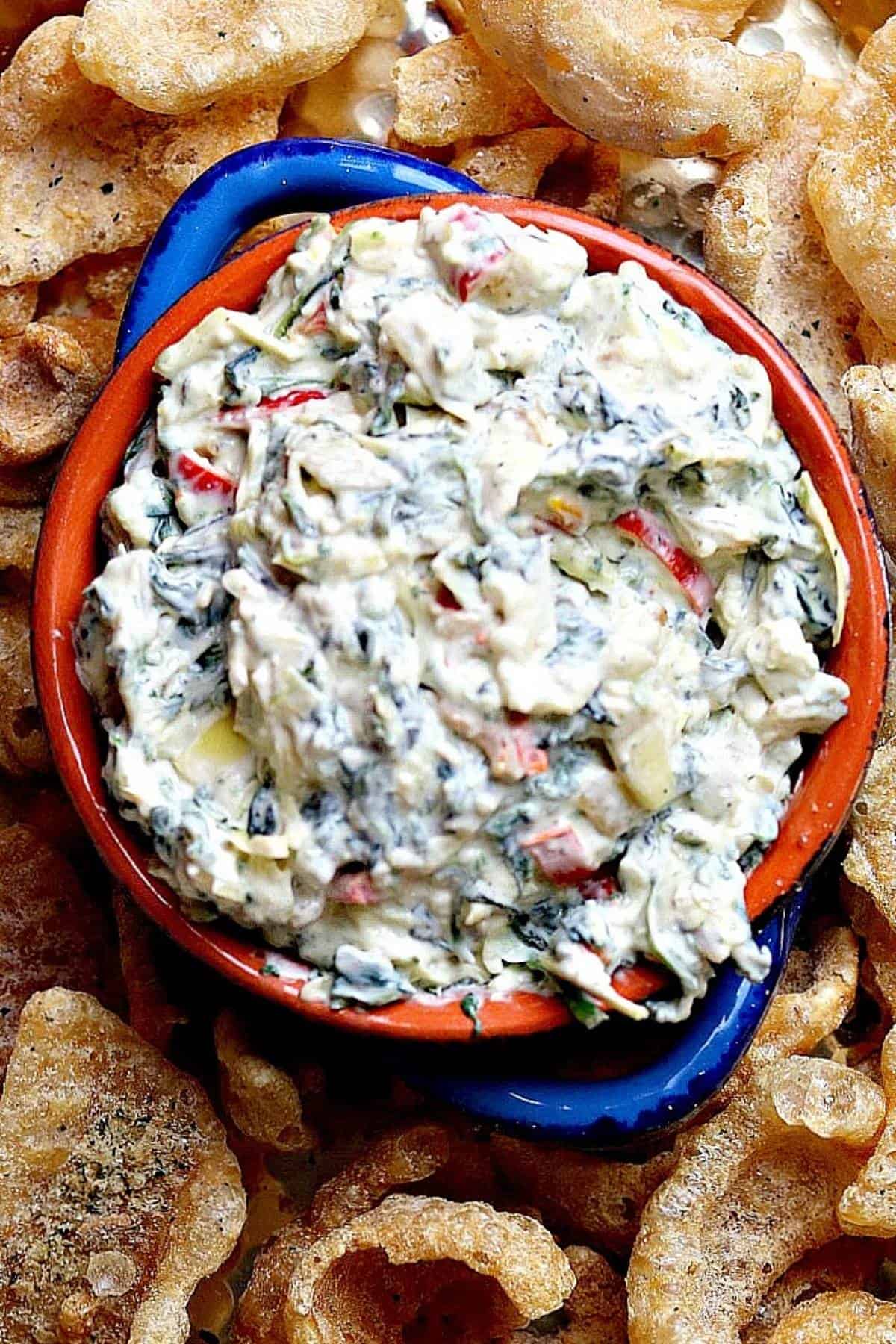 kale artichoke dip in a blue bowl surrounded by pork rinds