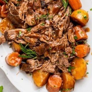 slow cooker pot roast on a plate with tender shredded beef chuck roast, gravy, carrots and potatoes, topped with rosemary and thyme
