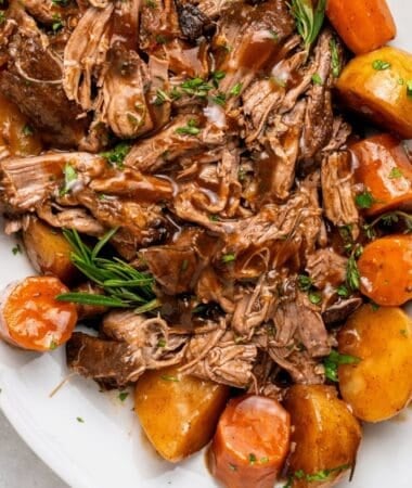 slow cooker pot roast on a plate with tender shredded beef chuck roast, gravy, carrots and potatoes, topped with rosemary and thyme
